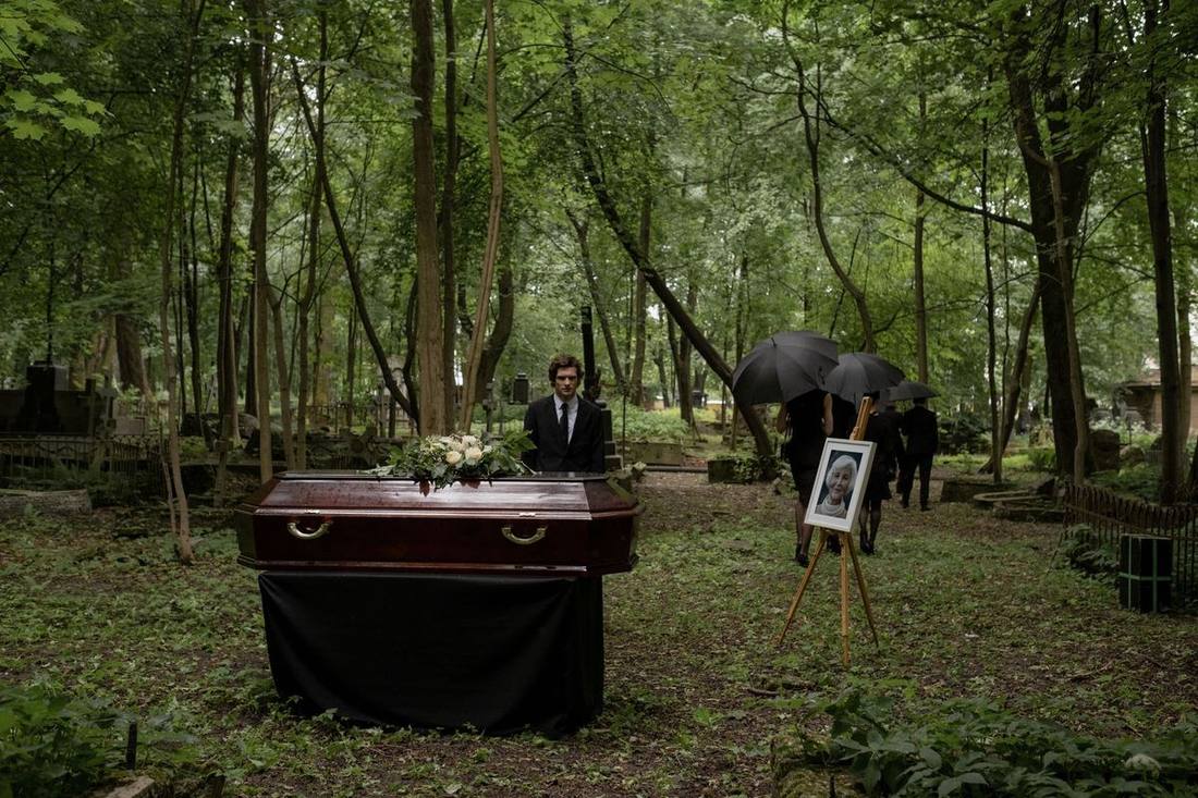 A chauffeur holding an umbrella for an elderly mourner at a cemetery road beside a black sedan