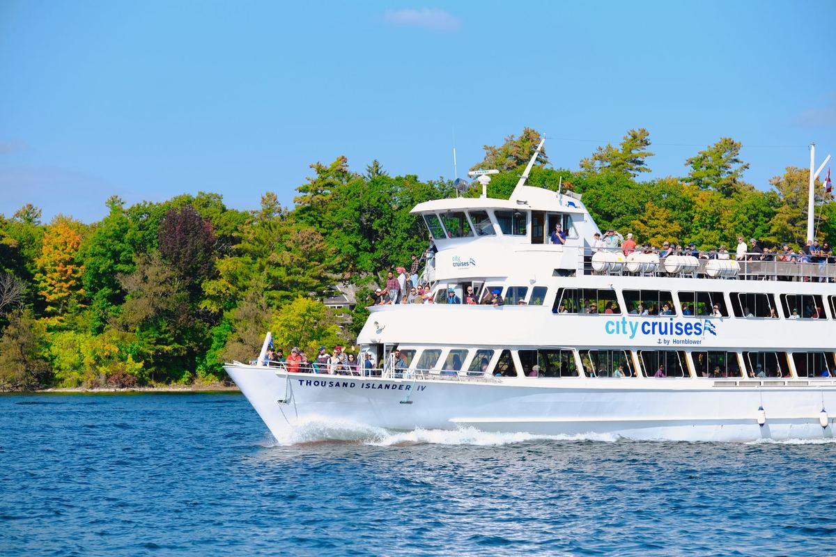Passengers boarding a Thousand Islands cruise boat at the Gananoque town dock with a black sedan parked nearby