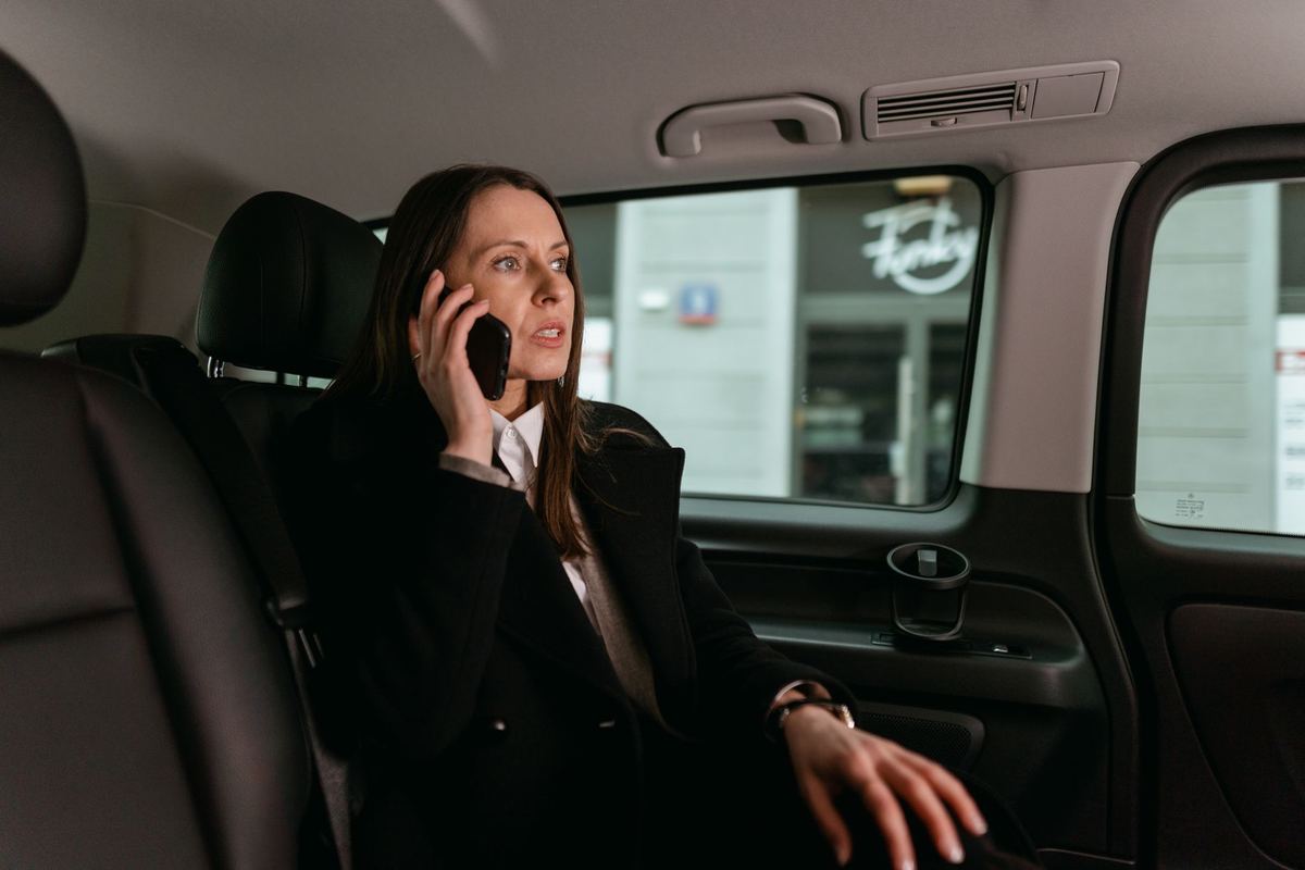 A female chauffeur in a dark suit holding a black SUV's rear door open for a passenger outside a hotel